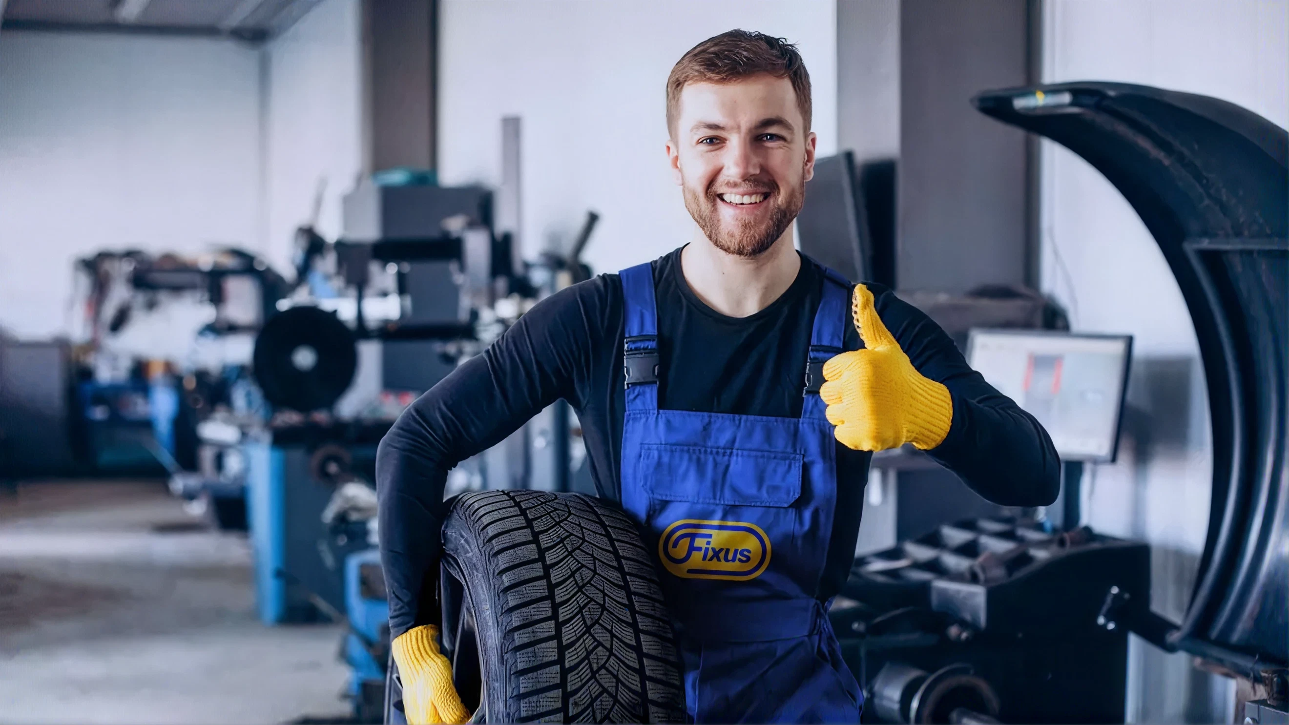 Hymyilevä mekaanikko pitelee auton talvirengasta ja näyttää peukkua korjaamolla